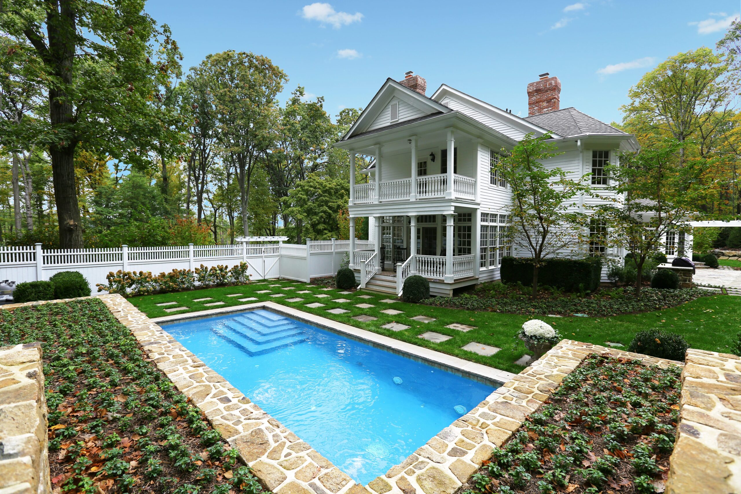 A beautiful two-story white house with a columned porch and a second-story balcony. In the foreground, a swimming pool is surrounded by a well-manicured lawn with stepping stones leading to the house. The pool is bordered by a stone retaining wall with low-growing plants. A white picket fence runs along the left side of the property, separating it from a wooded area.