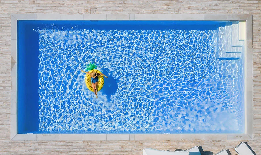 An aerial shot of a woman floating on a large, inflatable yellow pineapple raft in a rectangular swimming pool. The pool has a bright blue interior and is surrounded by a light-colored, tiled patio. Steps are visible in the corner of the pool.