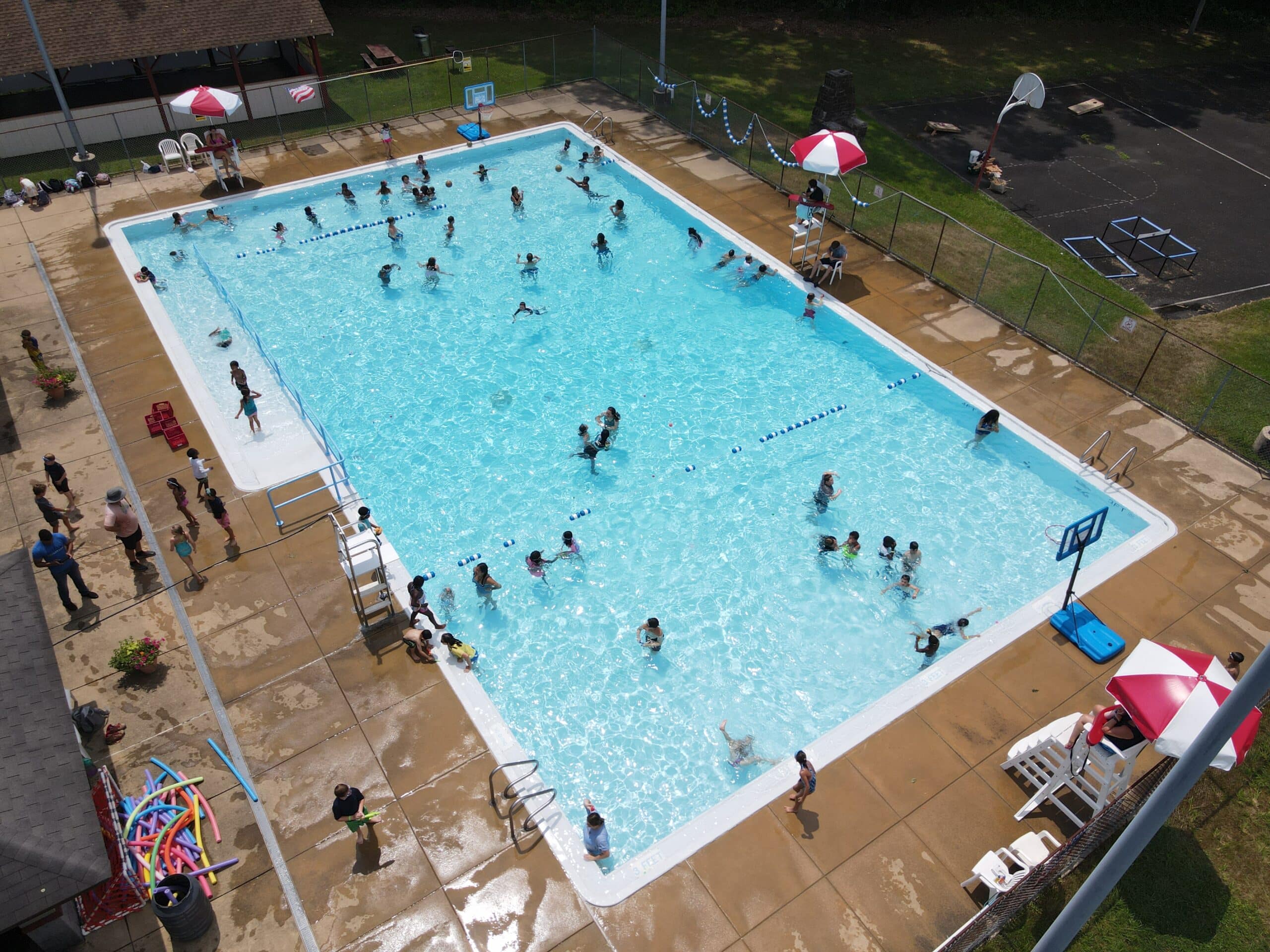 An aerial view of a rectangular community swimming pool on a sunny day. Many people, including adults and children, are swimming and playing in the clear blue water. A few people are sitting or standing on the concrete deck around the pool. There are chairs, umbrellas, and a basketball hoop near the edges of the pool.