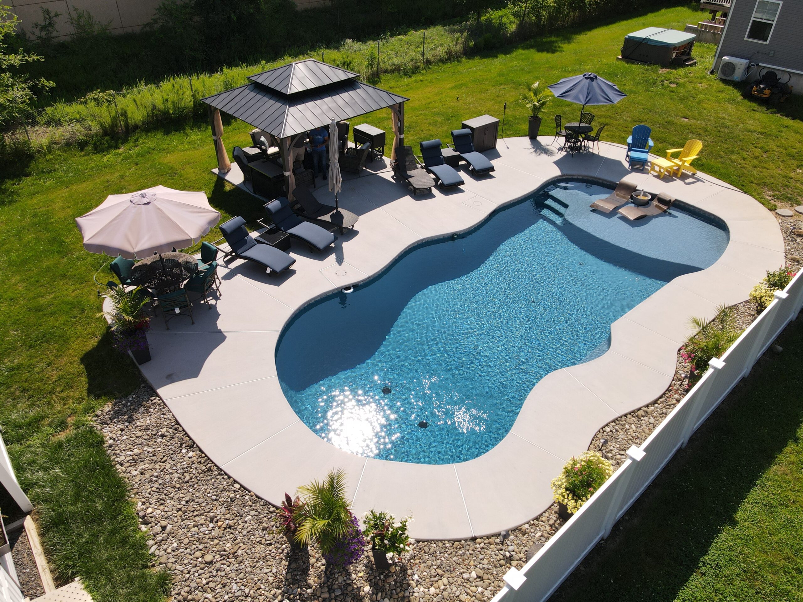 An aerial view of a backyard swimming pool with a unique, curvy shape and a sun shelf at one end. The pool has a dark blue interior and a light-colored concrete deck. On the sun shelf, there are two lounge chairs, and in the background, a few colorful Adirondack-style chairs are visible under a blue umbrella.