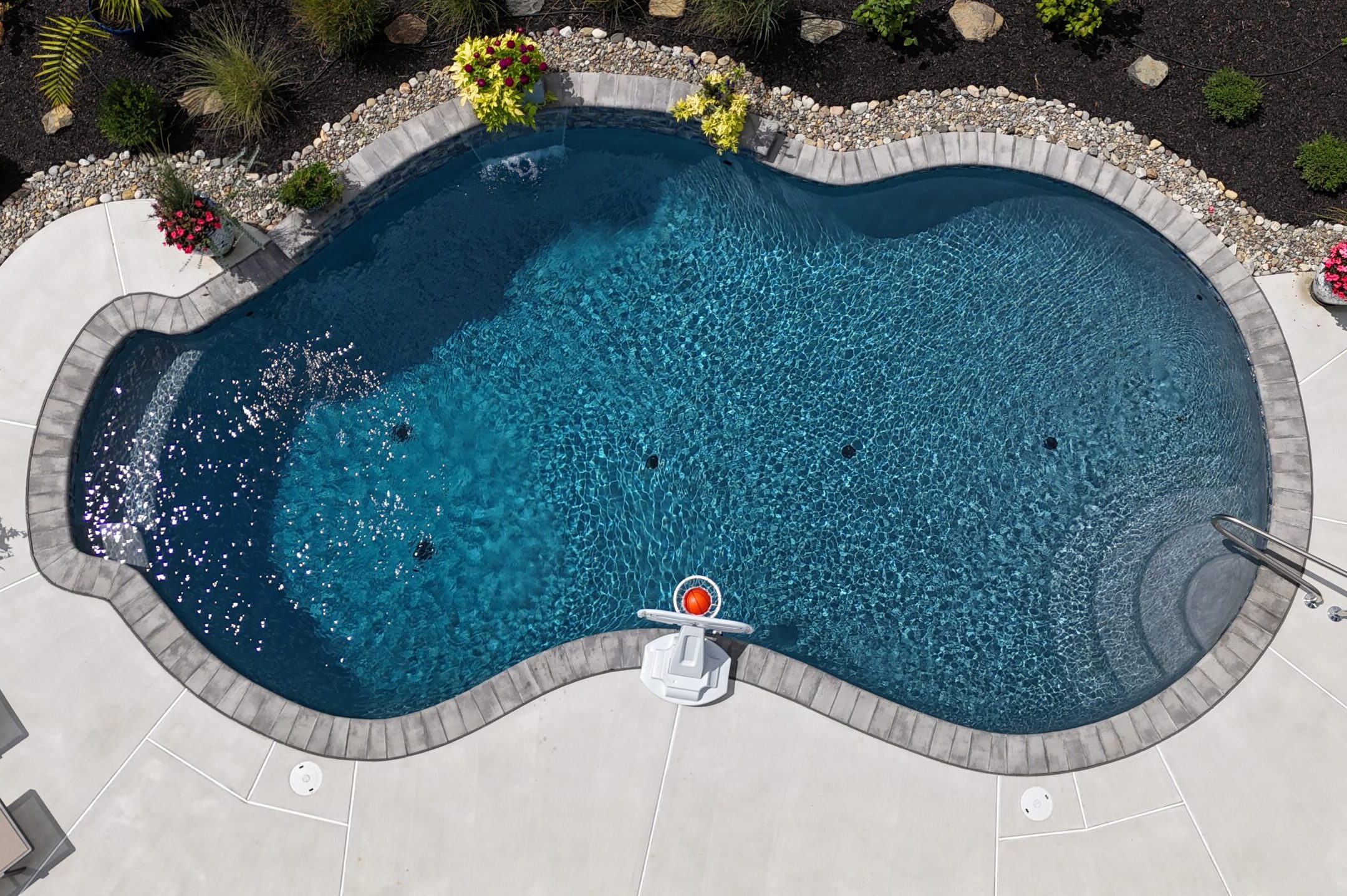 An aerial view of a custom-shaped swimming pool with a dark blue interior. The pool has a curved, irregular shape and is surrounded by a light-colored concrete deck with a decorative border of gray stones. At the top of the image, there are planters with red and yellow flowers, and the pool's edge is lined with river rocks and dark mulch. A stainless steel hand railing with steps is also visible on the right side. The water is calm and clear, showing a gradient of color from dark to light blue in the shallow end.
