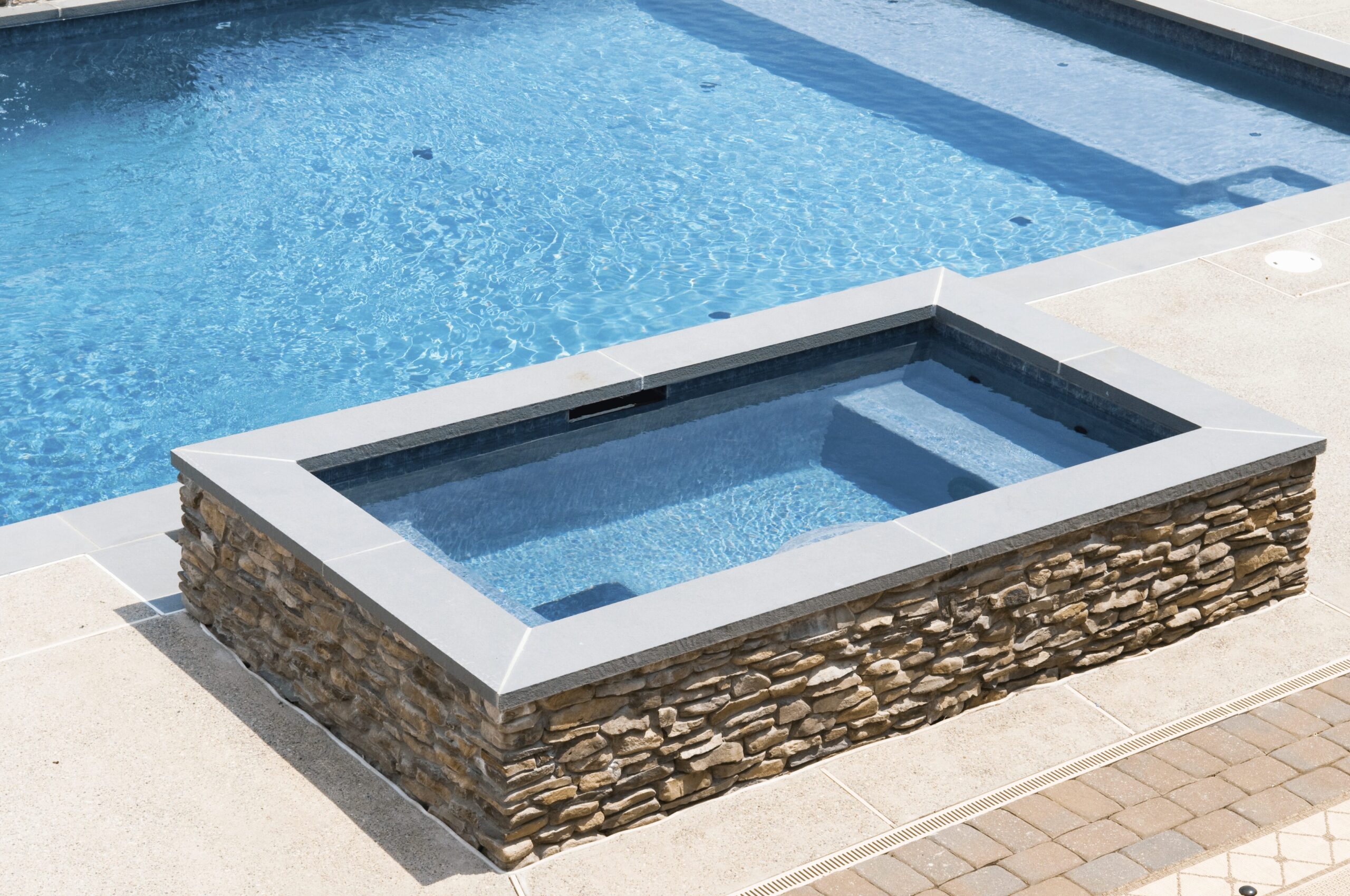 A rectangular, raised hot tub with a stone base and gray coping is next to a swimming pool. The water in the hot tub is a light blue, and steps are visible leading into it. The hot tub and pool are surrounded by a beige concrete patio.