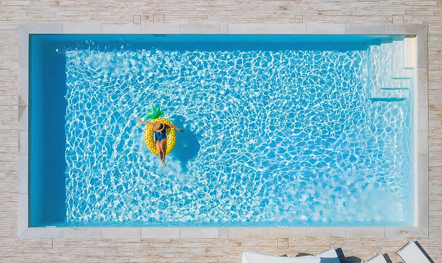 An aerial shot of a woman floating on a large, inflatable yellow pineapple raft in a rectangular swimming pool. The pool has a light blue interior and is surrounded by a light-colored, tiled patio. Steps are visible in the corner of the pool.