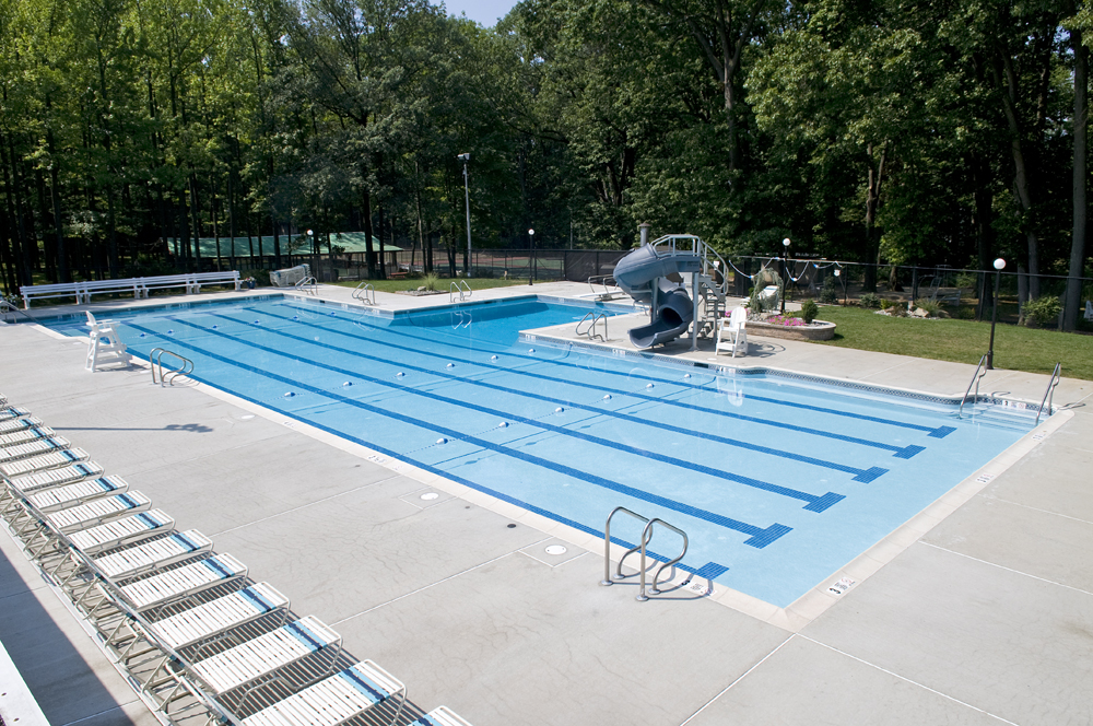 A bright, outdoor community swimming pool with several lanes marked with blue lines. A short, enclosed water slide is located at the far end of the pool, which is surrounded by a concrete deck and a black fence. Tall trees form a dense green backdrop behind the fence.