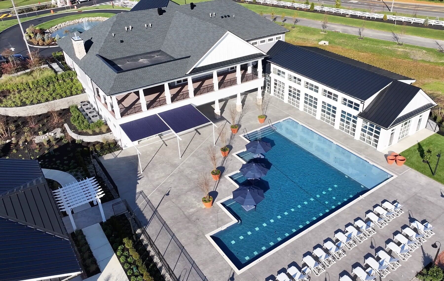 An aerial shot of a rectangular community swimming pool with a unique, stepped-out design on one side. The pool has a light blue interior and is surrounded by a light grey concrete deck. The deck features several rows of lounge chairs and three large, dark blue umbrellas for shade. A walkway with a white pergola leads to a building on the right.